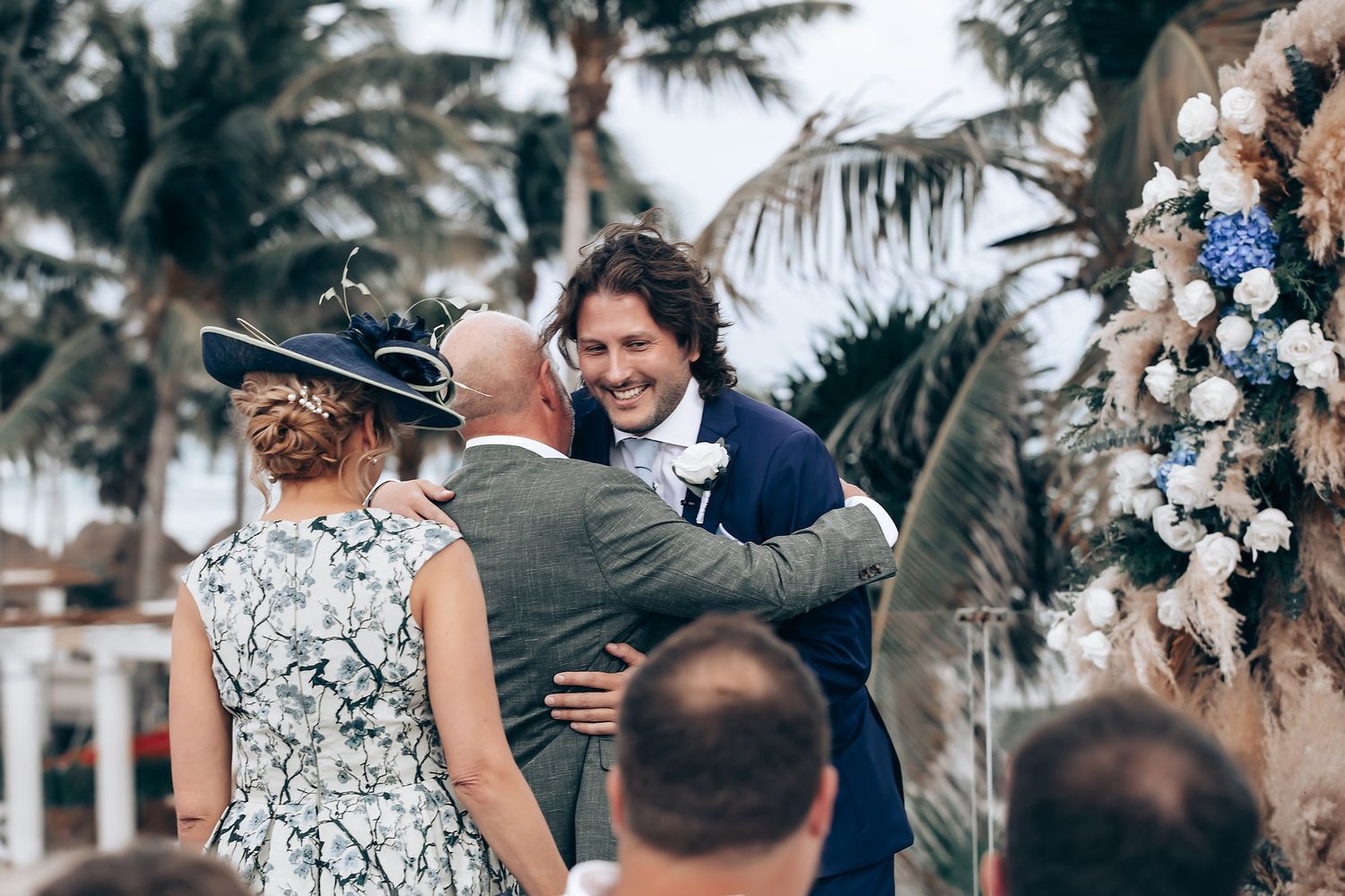 An outdoor wedding ceremony under palm trees with guests gathered around a floral arch.