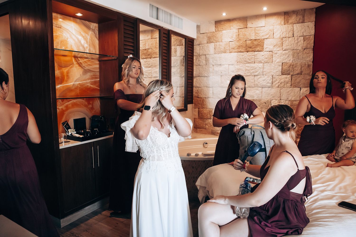 Wedding preparation scene with bridesmaids in burgundy dresses helping with final touches.