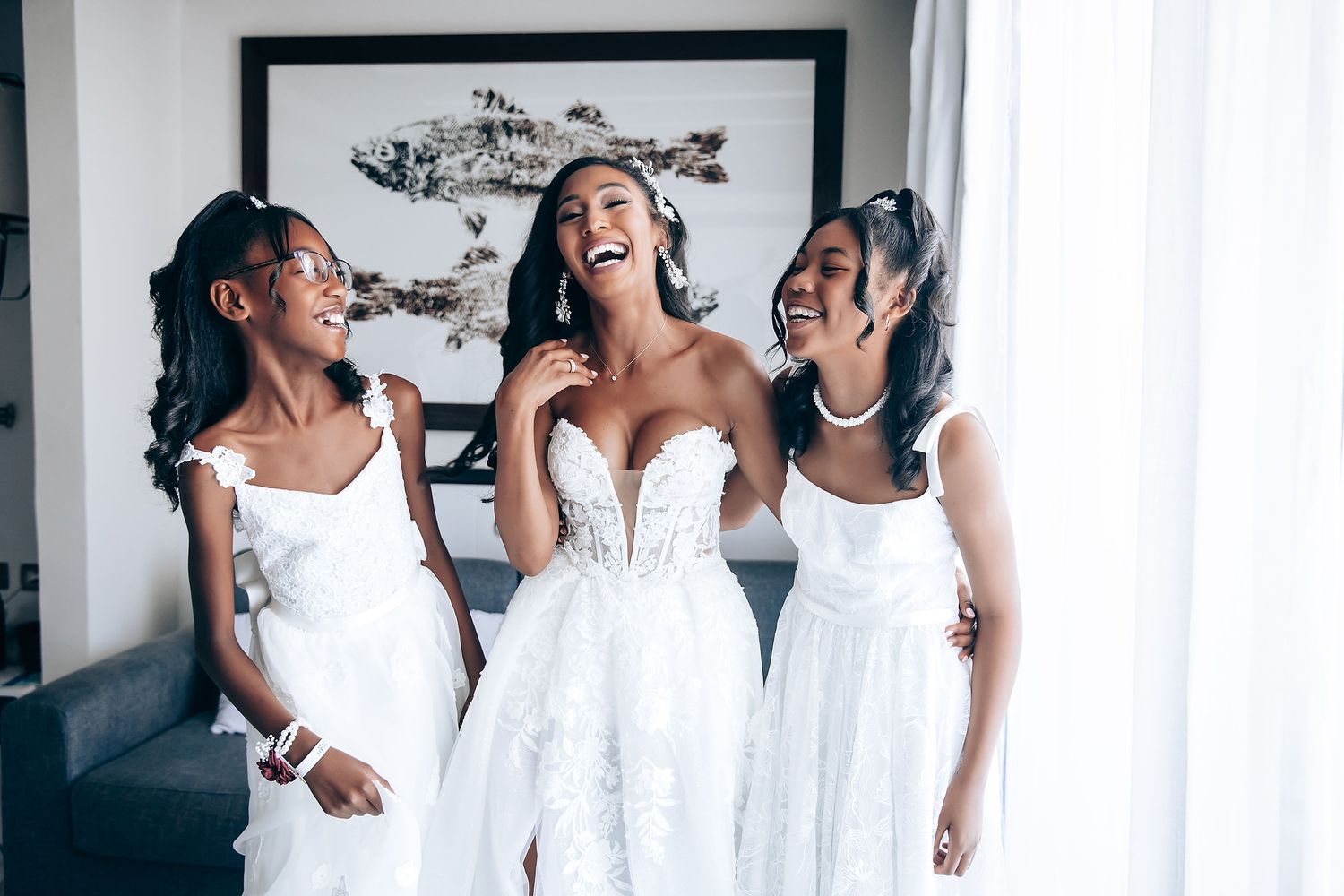 Three bridesmaids in white dresses share joyful laughter during wedding preparations.