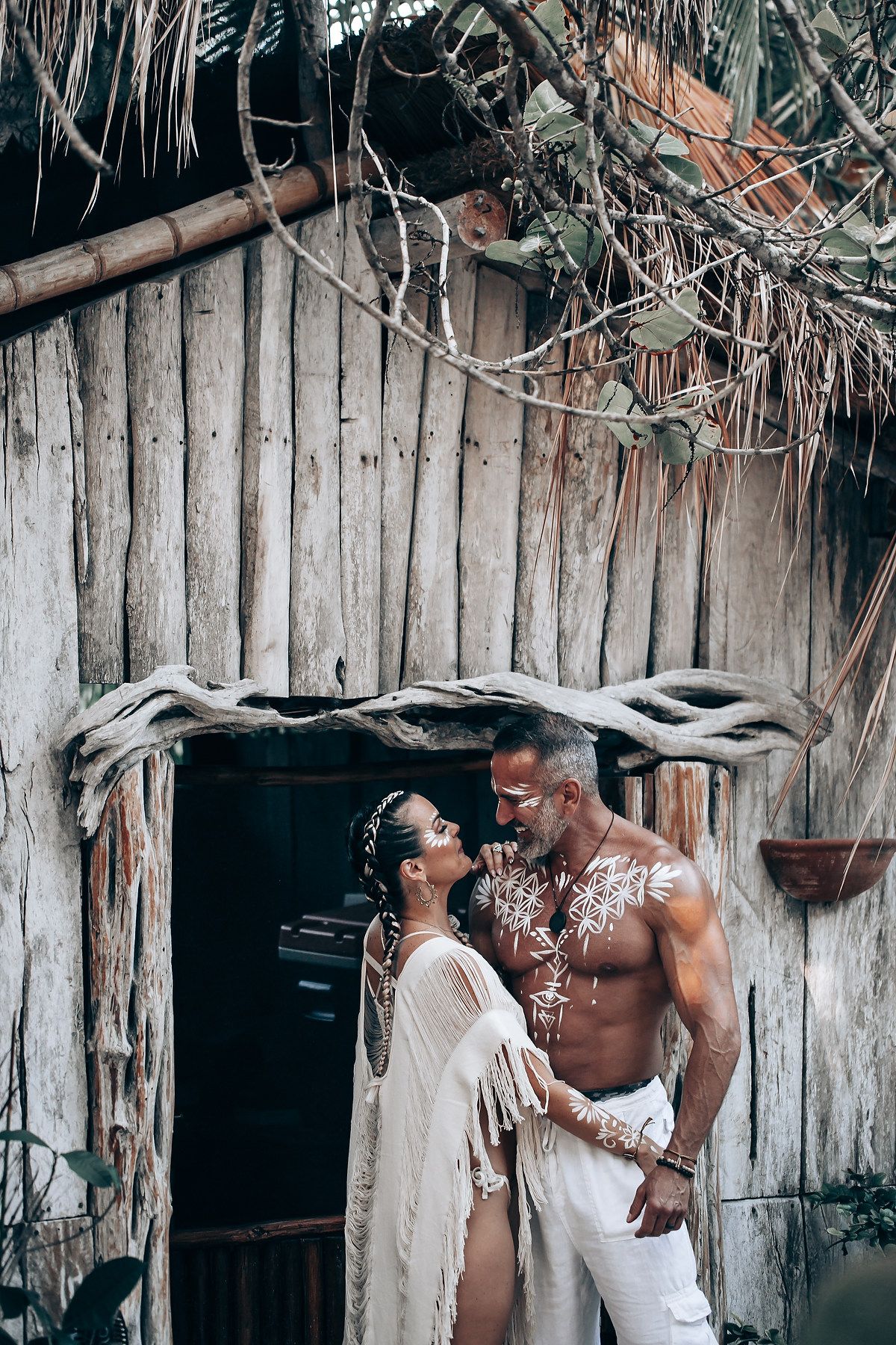 A couple poses intimately against a rustic wooden beach hut with tropical foliage.