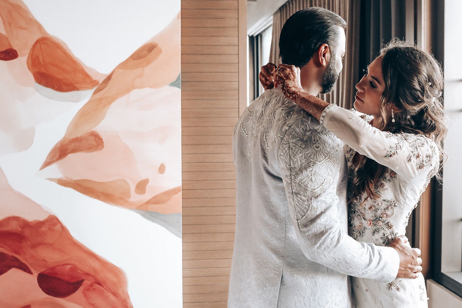 Couple in elegant white lace outfits embrace by window during romantic wedding photoshoot.