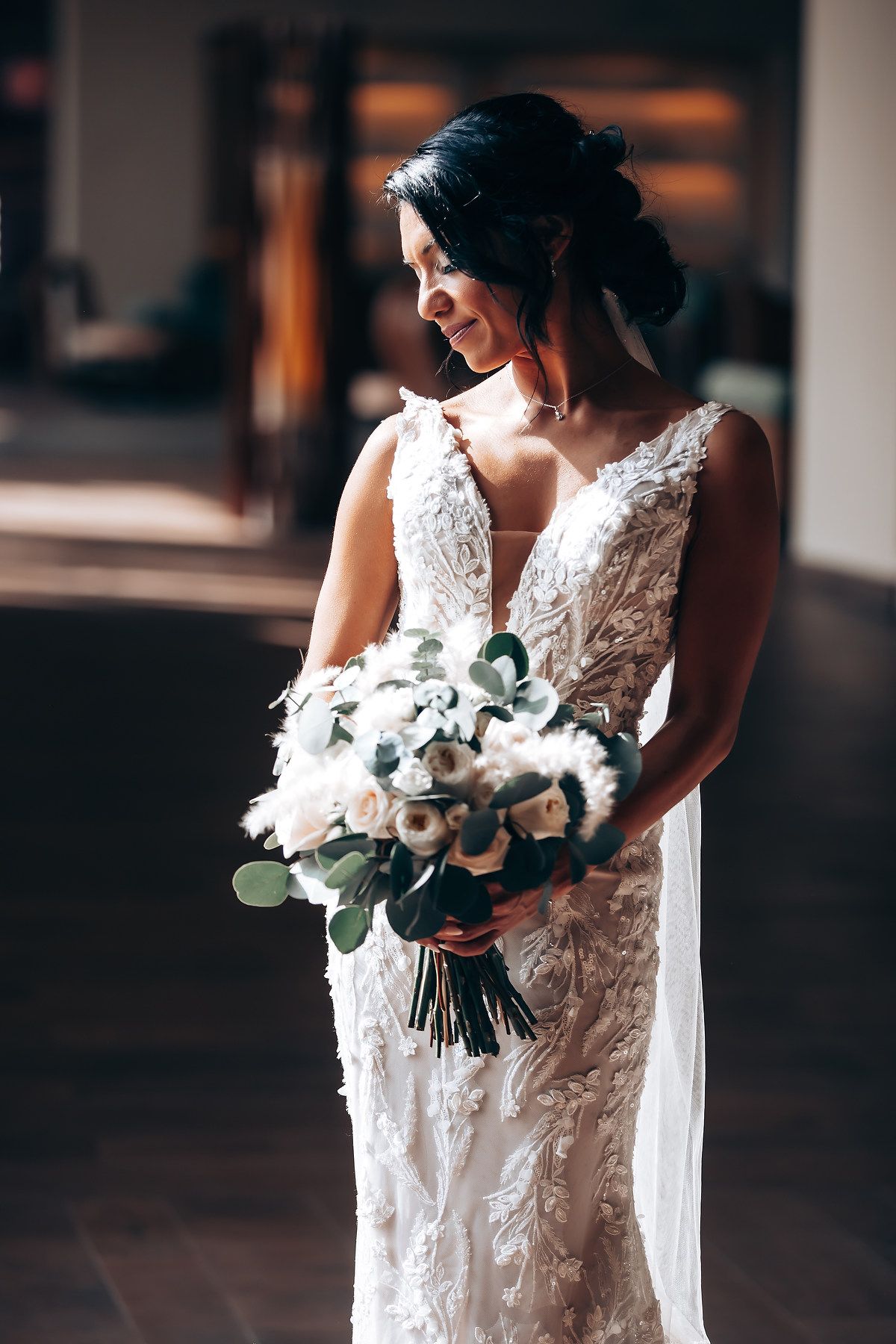 A bride in a detailed lace wedding dress holds an elegant white and green bouquet in an indoor setting.