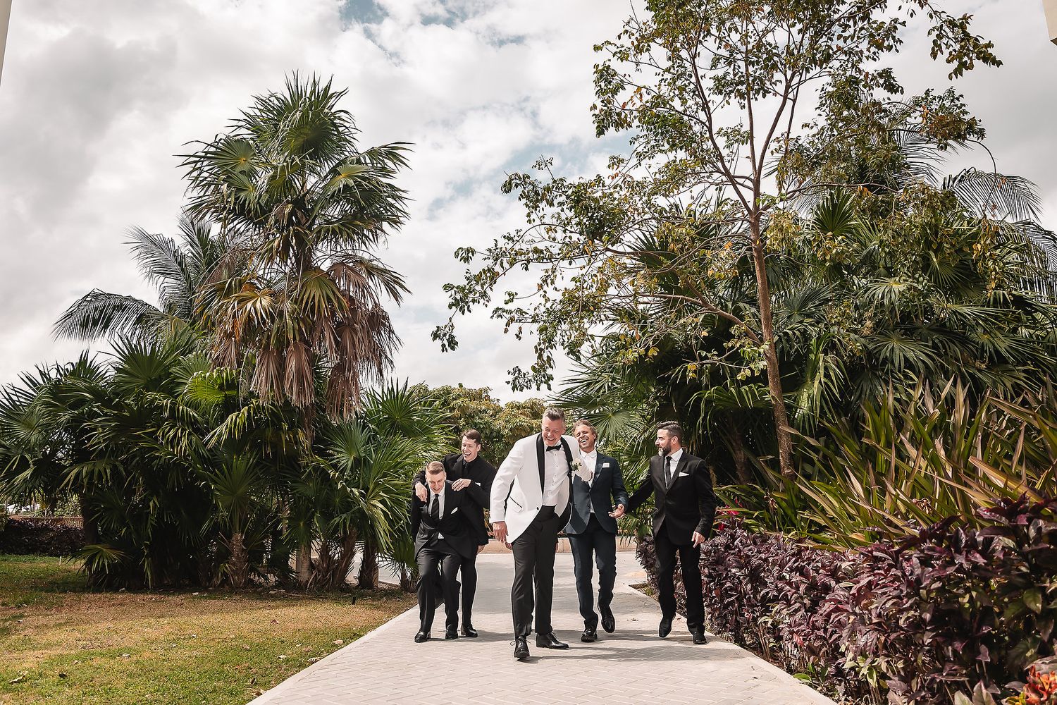 Wedding party in formal attire gathered outdoors with palm trees and cloudy sky in the background.