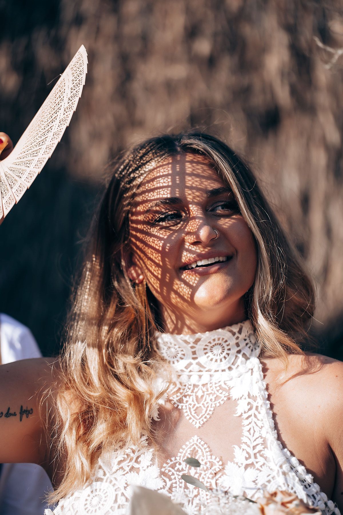 Woman in white lace dress laughs joyfully while sunlight creates dappled shadows across her face.