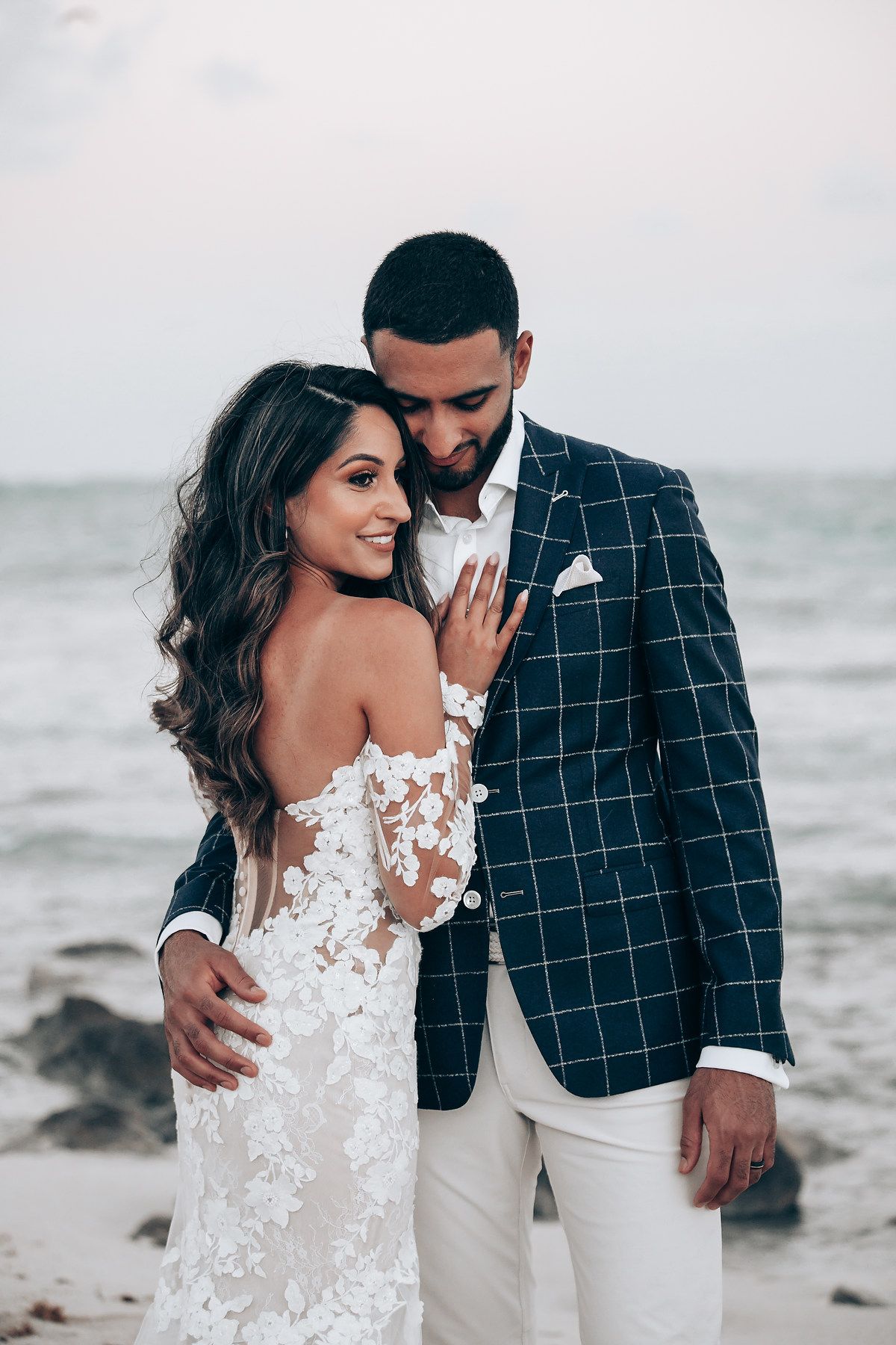 A couple poses on a beach at sunset, with bride in lace dress and groom in checkered blazer.