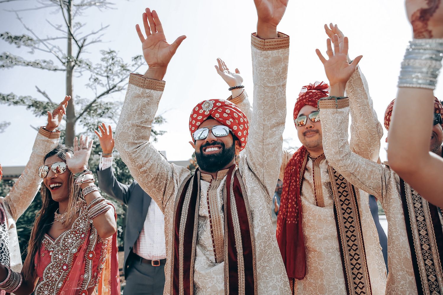 Group of wedding guests in traditional Indian attire raising hands in celebration.