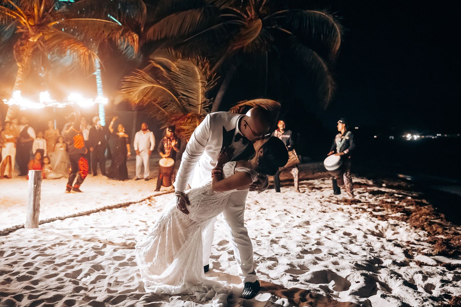 Couple performs romantic dance dip on beach during their wedding reception with fire performers nearby.