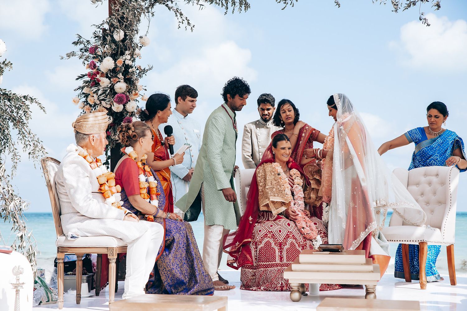 Wedding guests seated at a beachside ceremony under white canopy with floral decorations.