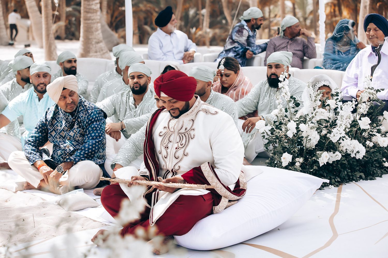 Traditional Indian wedding ceremony guests seated on white cushions under palm trees and floral decorations.