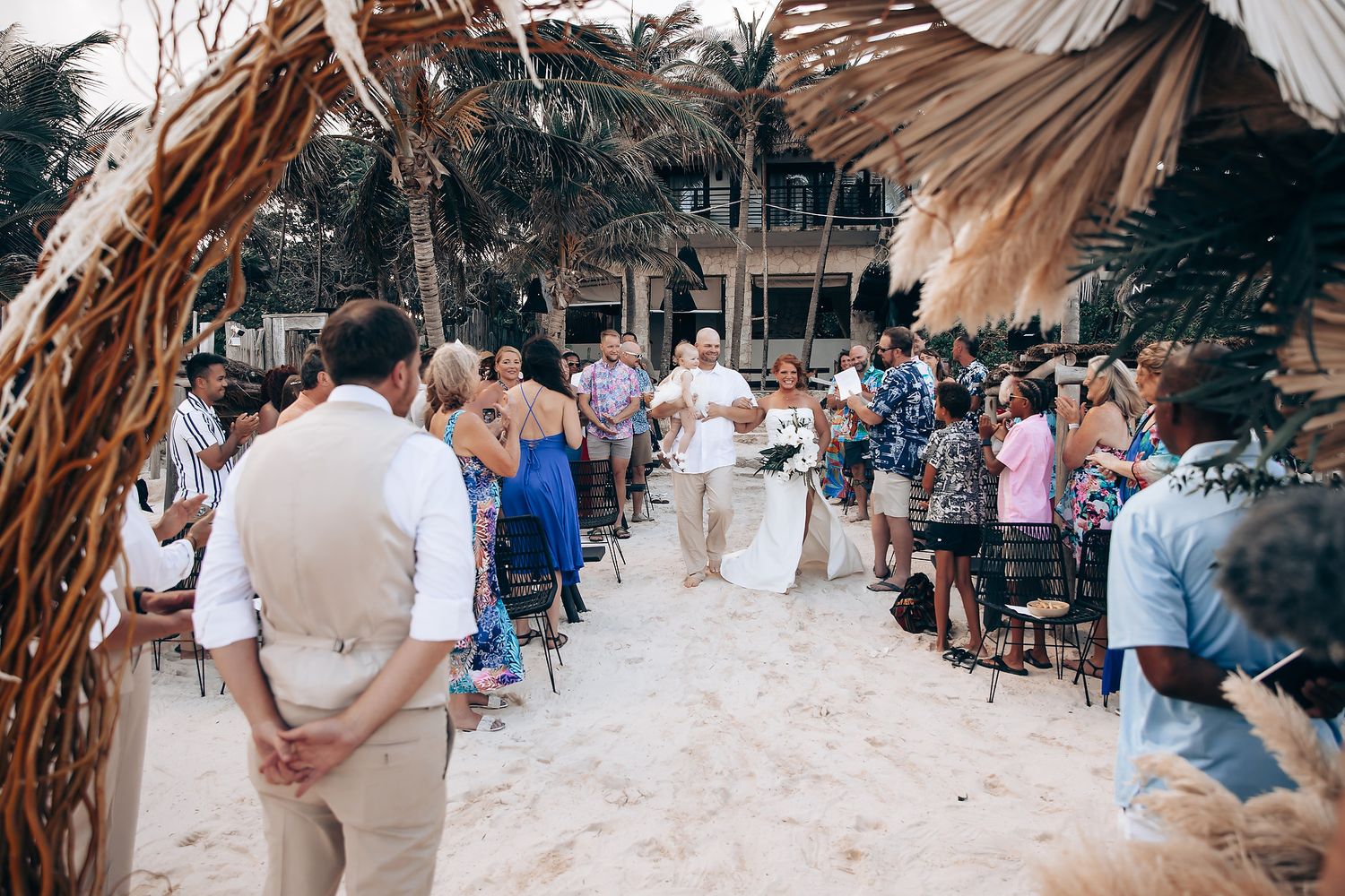 Beach wedding ceremony setup with guests seated under palm trees by the ocean.