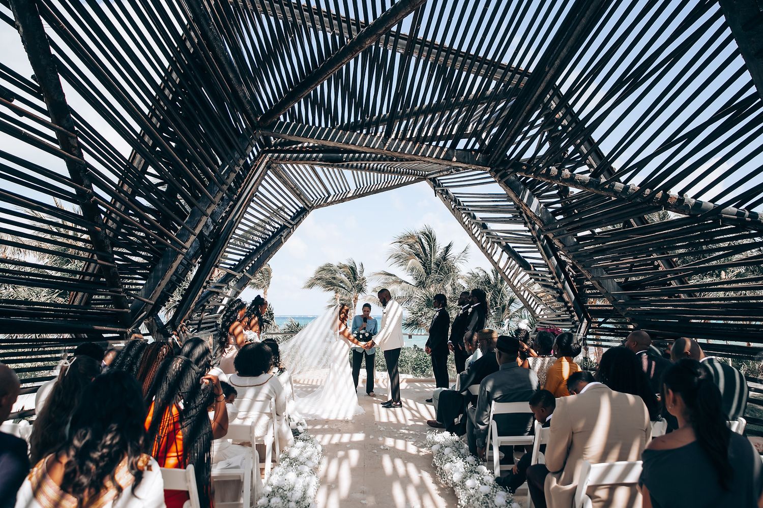 Outdoor wedding ceremony under a modern black geometric pergola with ocean and palm trees in background.