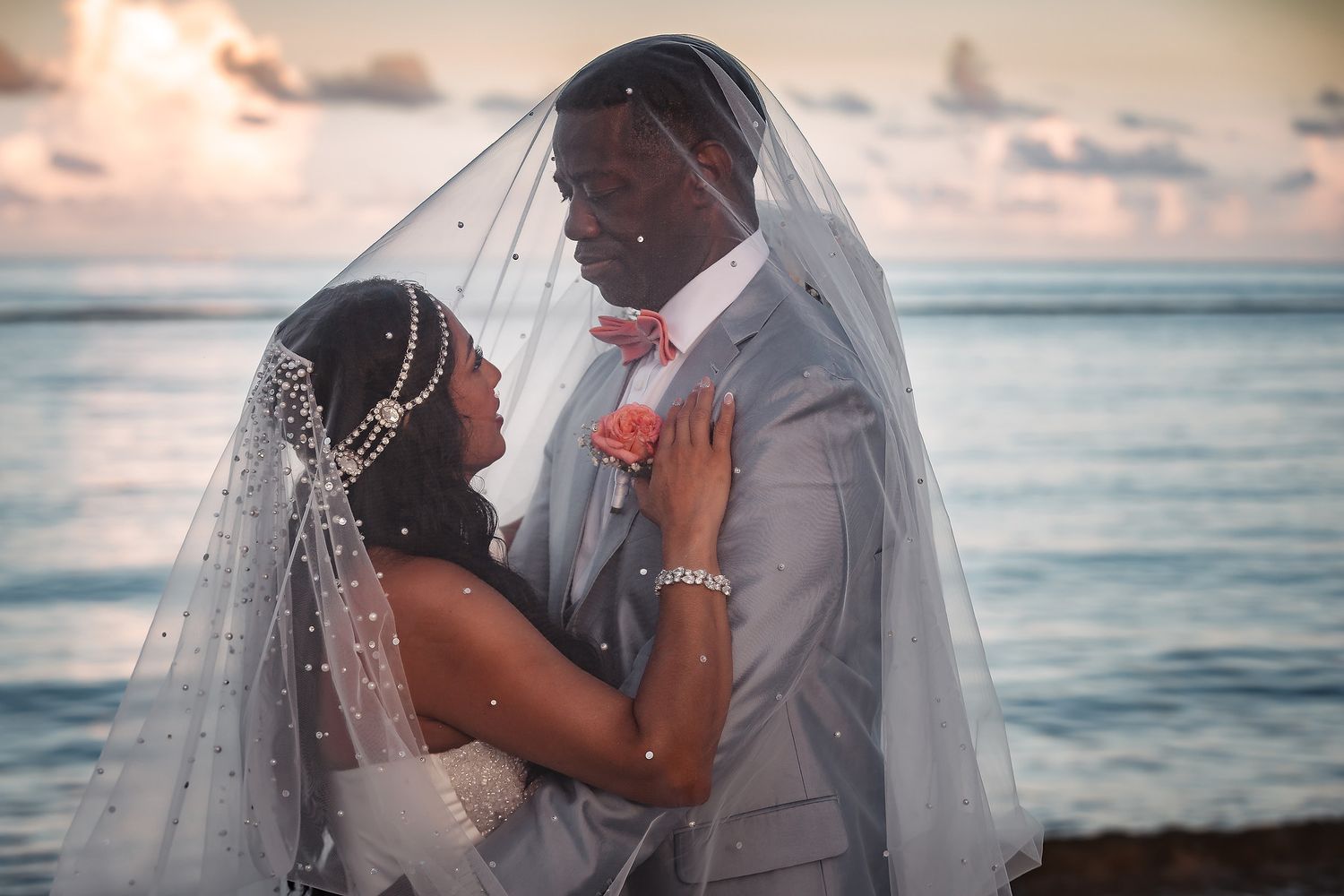 Romantic beach wedding portrait with flowing veil against sunset sky.