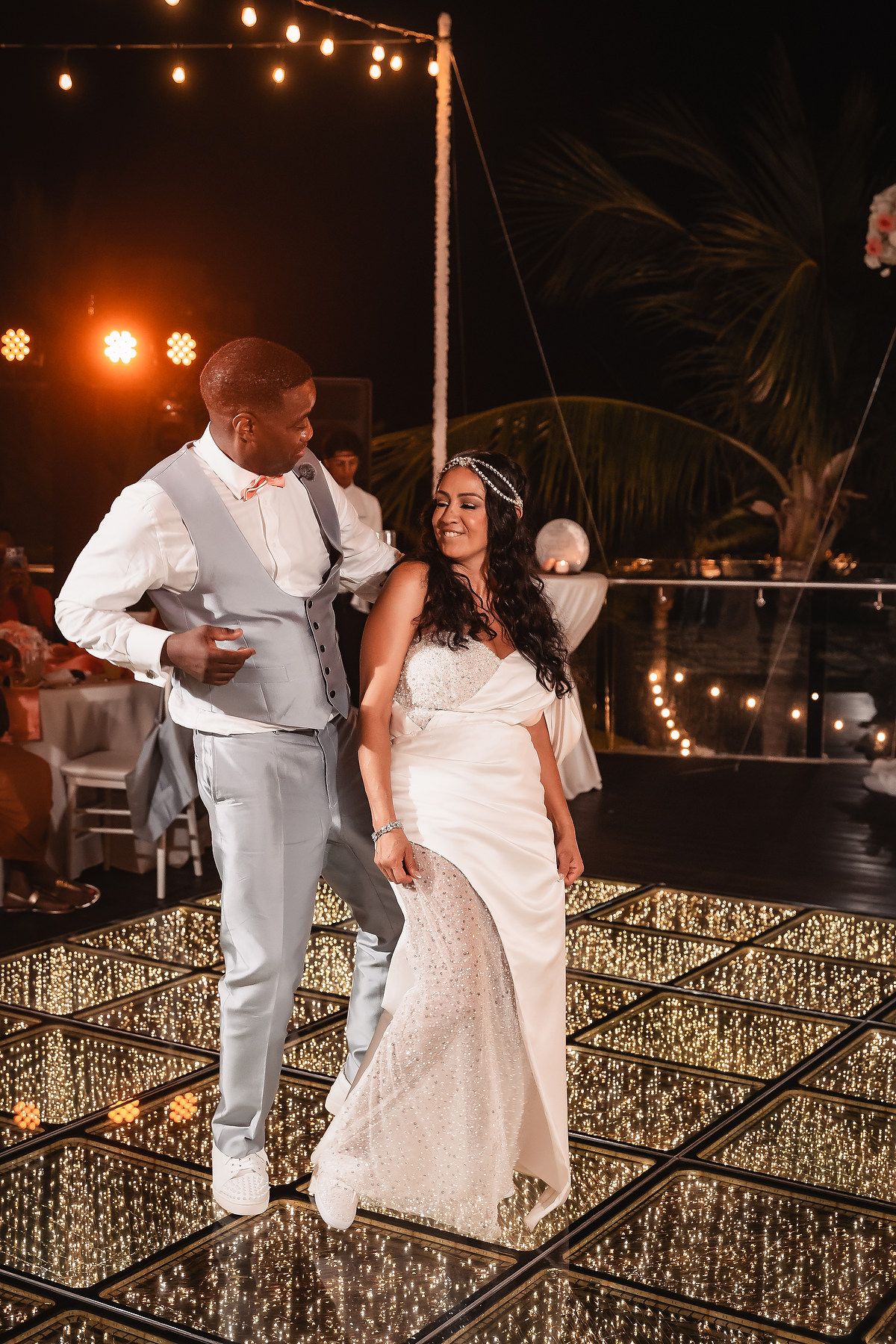 Couple dancing on a mirrored floor under string lights at an outdoor evening reception.