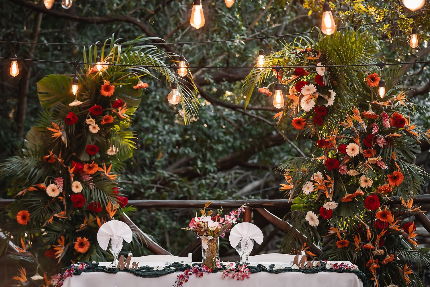 Garden wedding table setting with white linens and red floral arrangements illuminated by hanging lights.