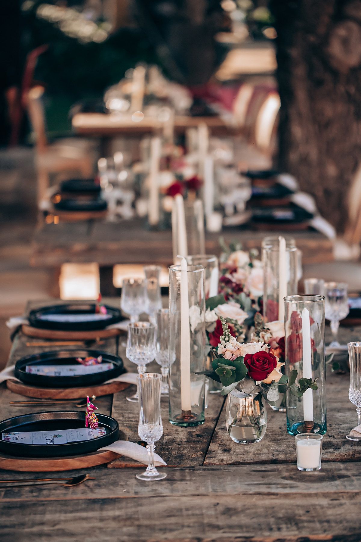 Rustic wedding table setting with navy blue plates, tall white candles and burgundy floral centerpieces on wooden table.