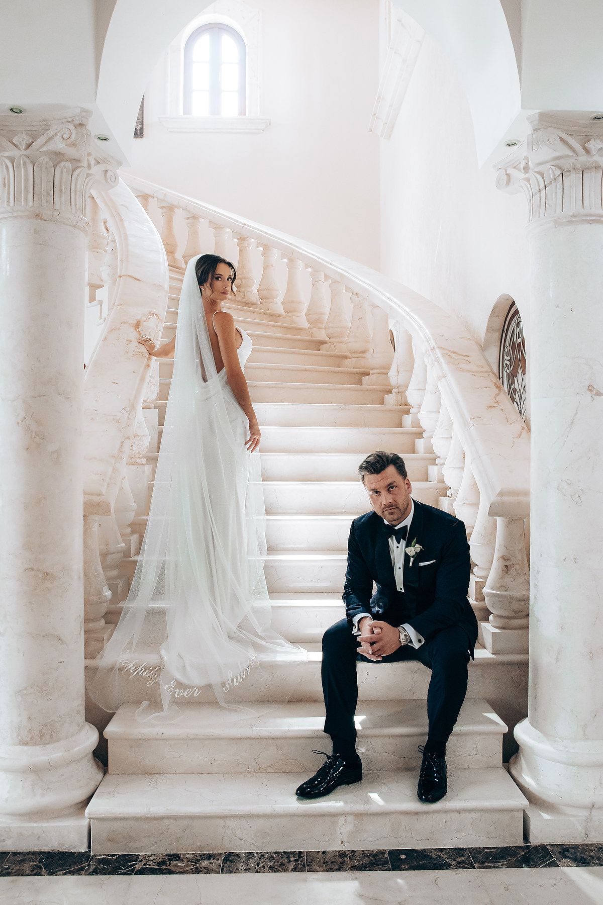 Bride and groom sit separately on elegant white marble staircase in wedding attire creating artistic composition.