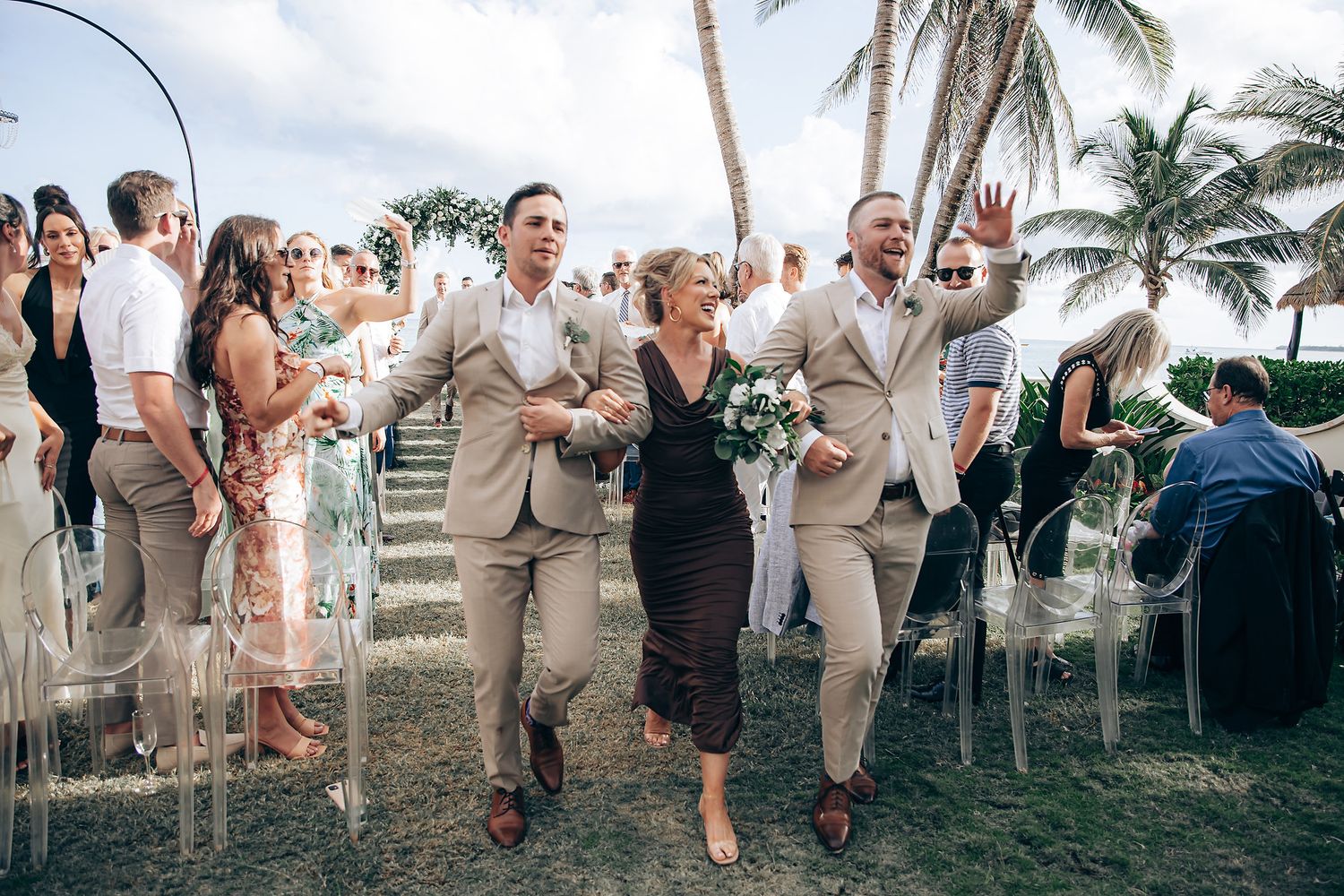 A group of wedding guests in beige and white attire pose together on a tropical beach with palm trees at sunset.