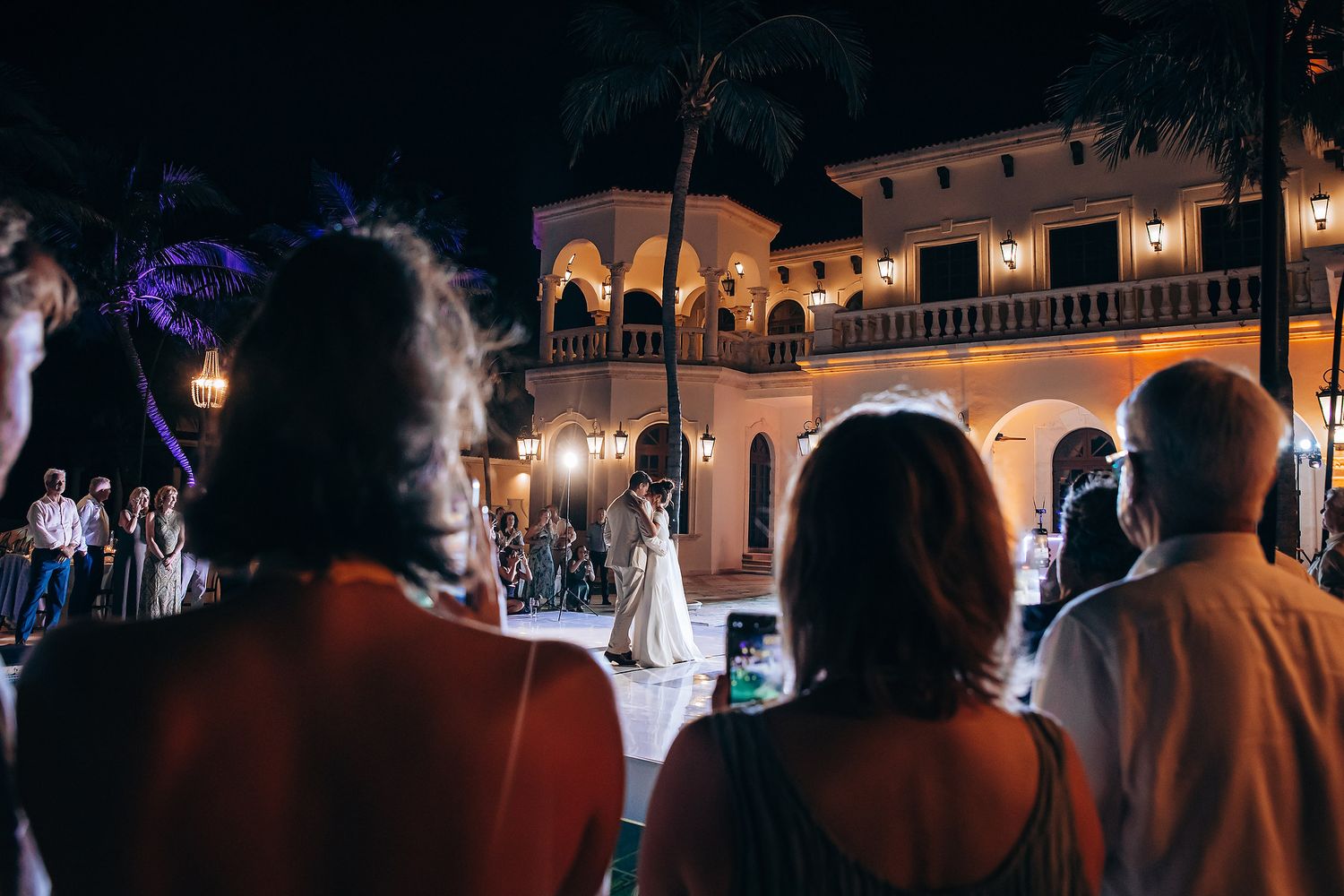 Silhouettes of people enjoying a nighttime event with colorful lights illuminating a building facade in the background.