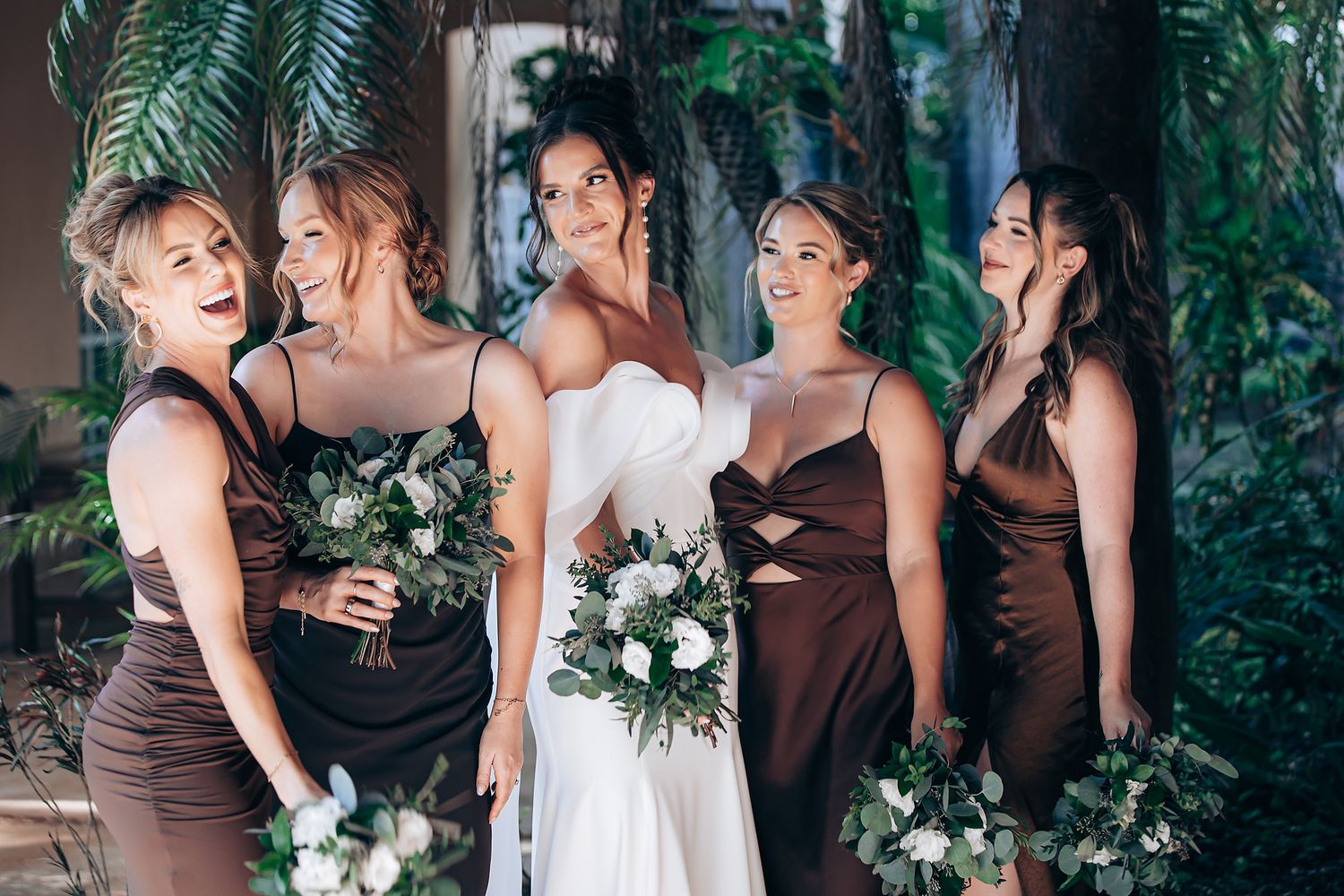 Wedding party members in brown and white dresses pose together against a lush green garden backdrop.