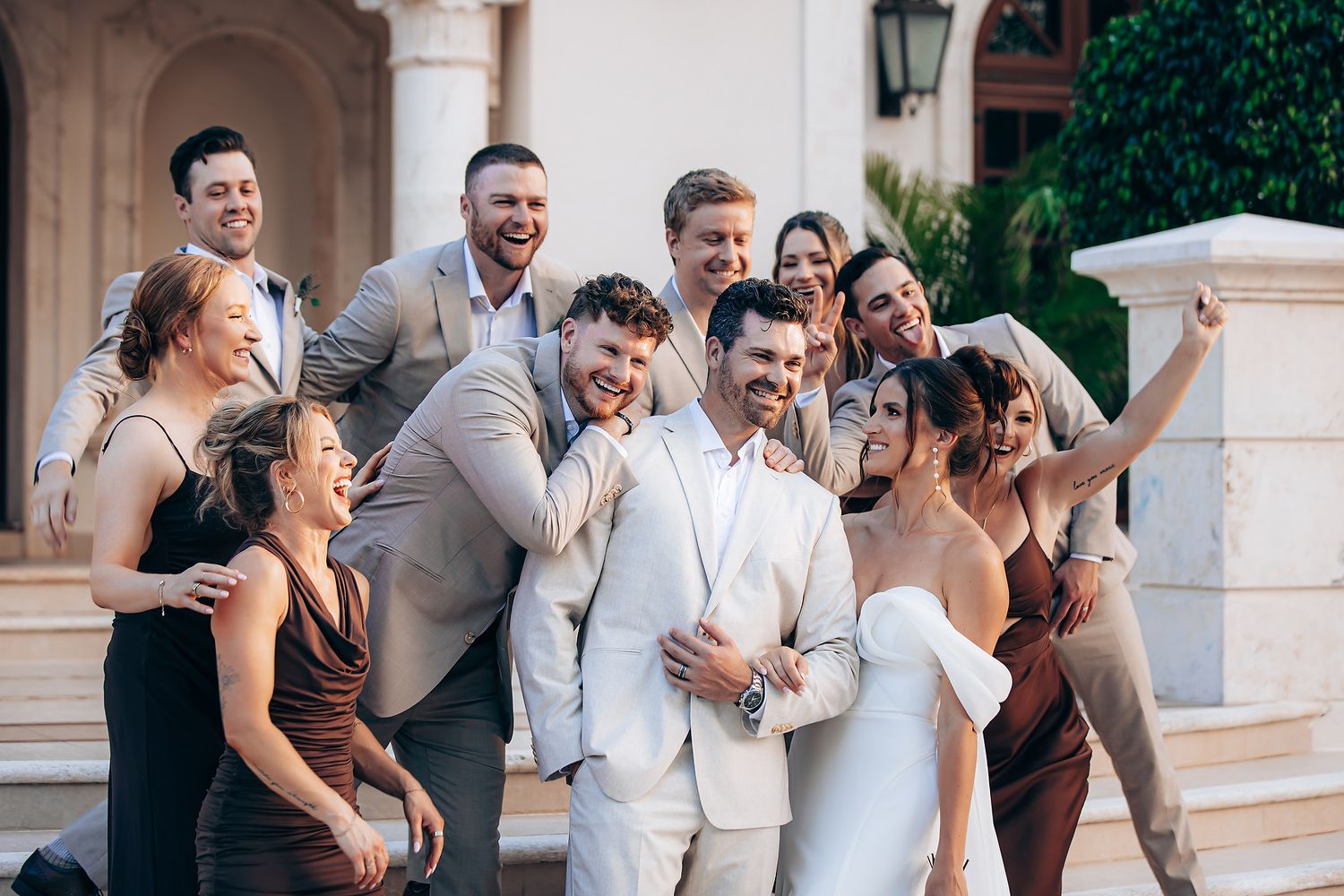 Wedding party in formal attire poses together outdoors at an elegant venue with stone columns and archways.