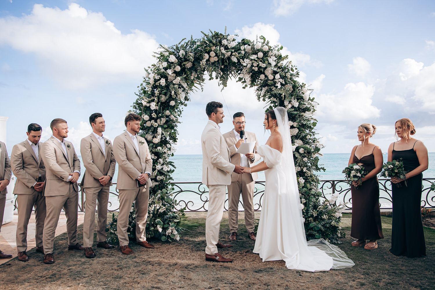 Wedding party poses outdoors under a floral arch during a beachside ceremony.