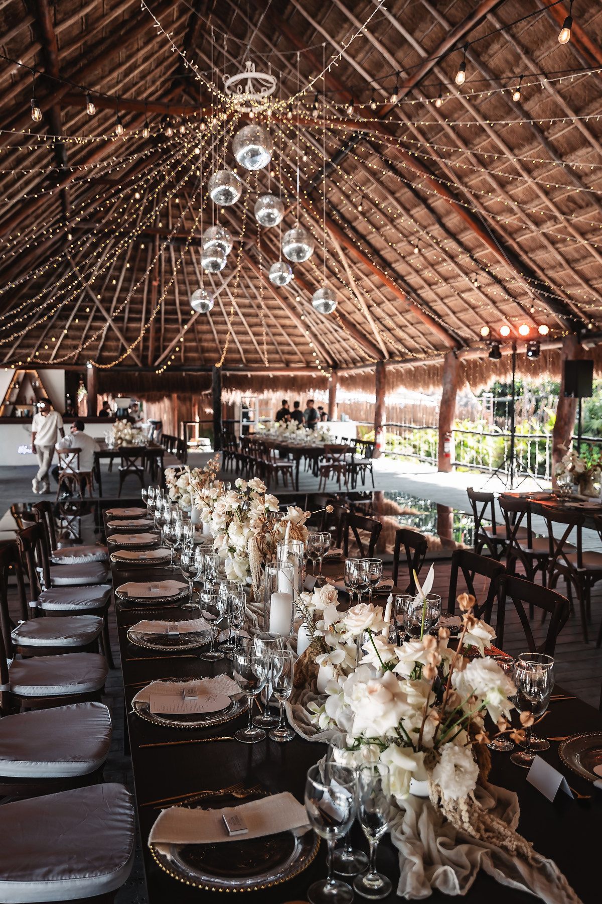 Elegant reception setup in thatched roof venue with hanging disco balls and white floral table decorations.