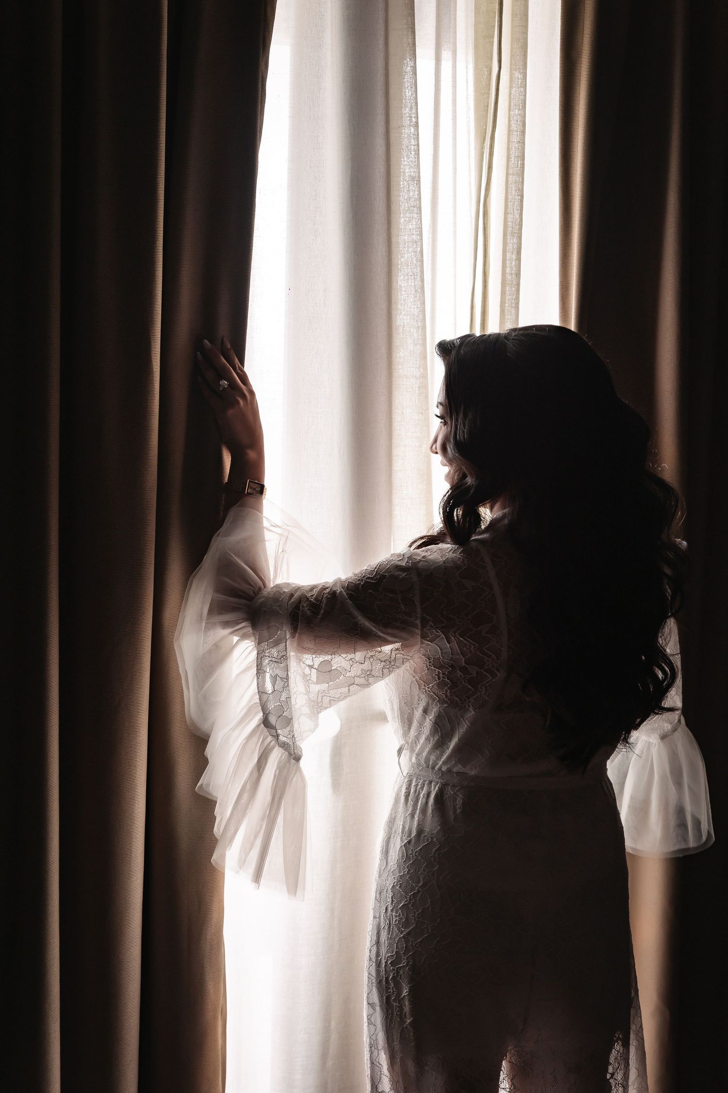 Silhouette of someone in white lace standing by a window with flowing curtains.