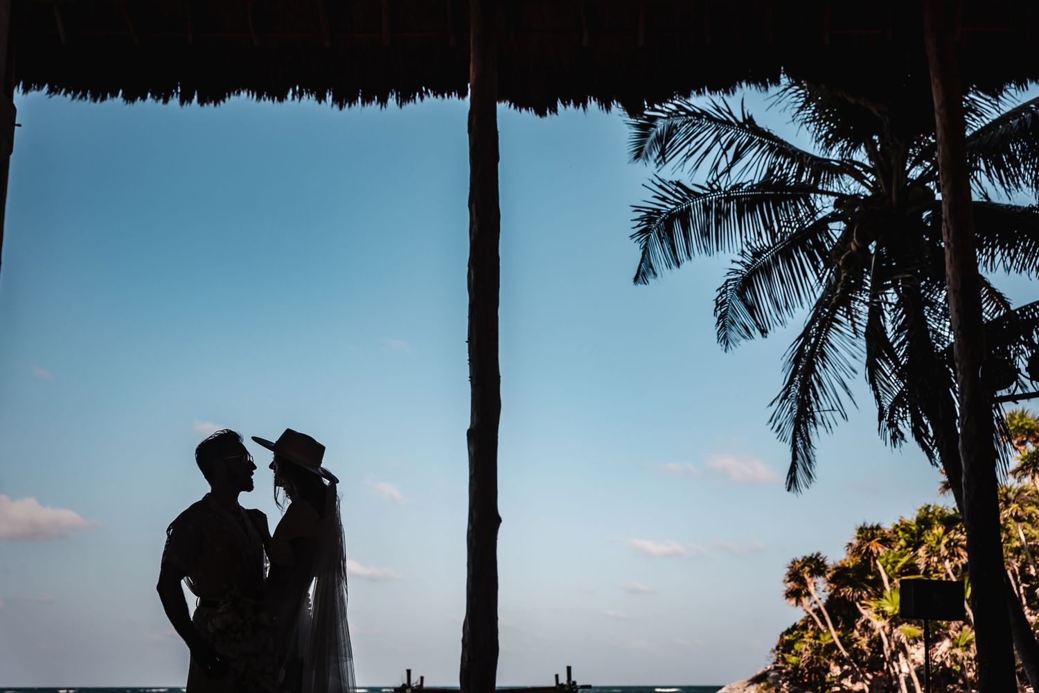 Silhouetted palm trees frame a romantic beachside wedding scene at sunset.
