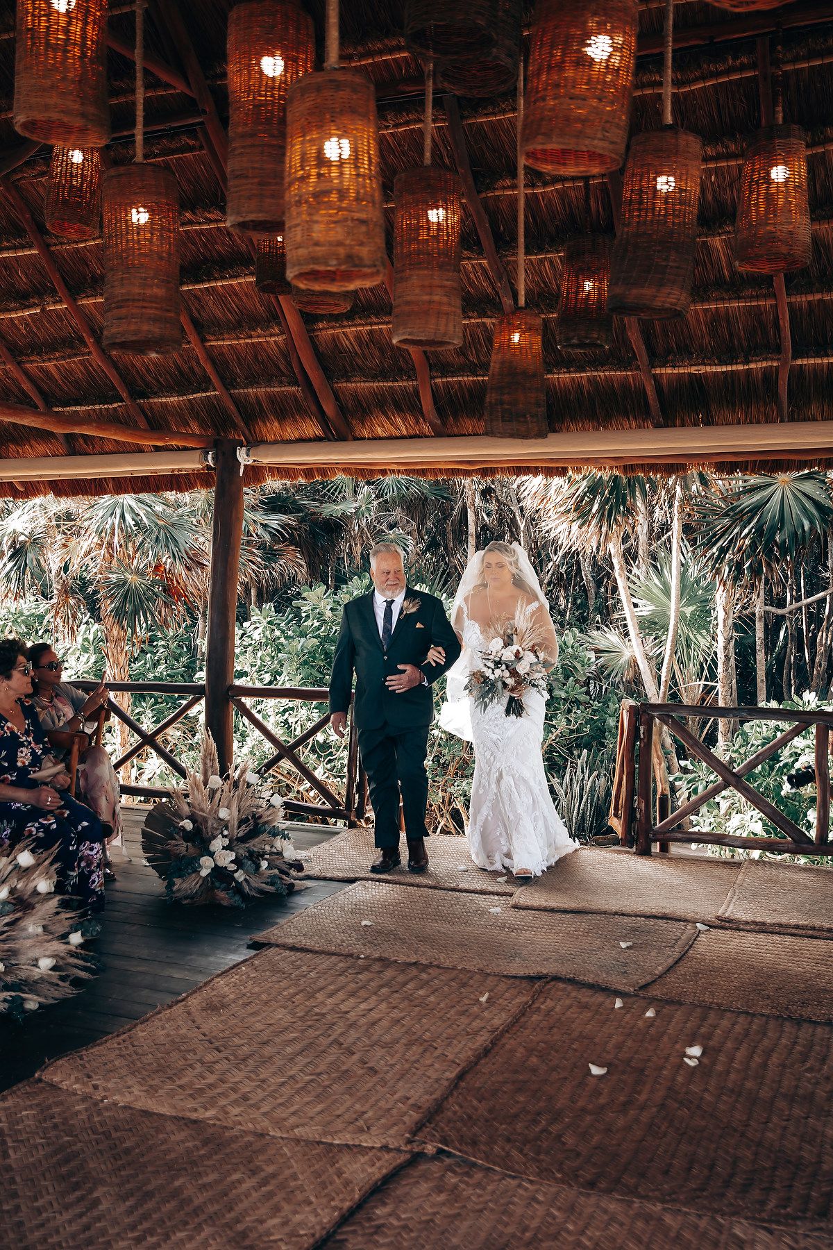Wedding ceremony in progress under a rustic thatched roof pavilion with hanging lights.
