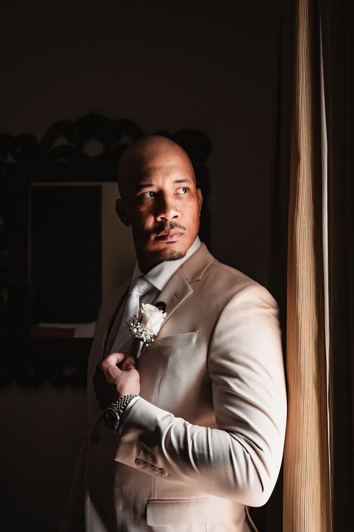 Portrait shots in dramatic lighting showing a groom in a cream suit with boutonniere against a dark background.