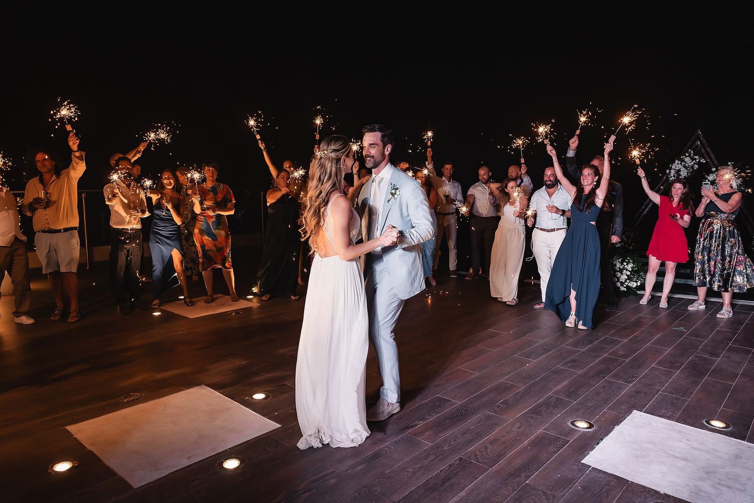 Wedding couple embraces on dance floor as guests surround them with sparklers at night.