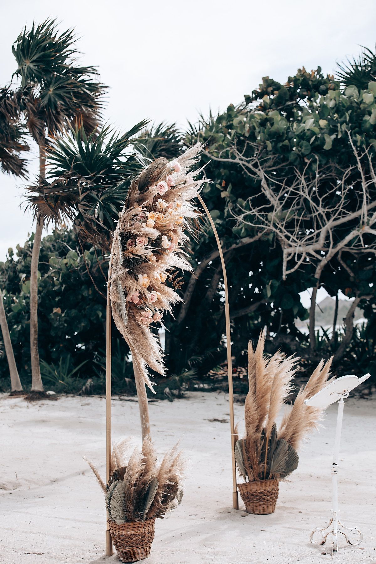 Pampas grass and palm trees decorate a tropical beach wedding ceremony setup with natural woven baskets.