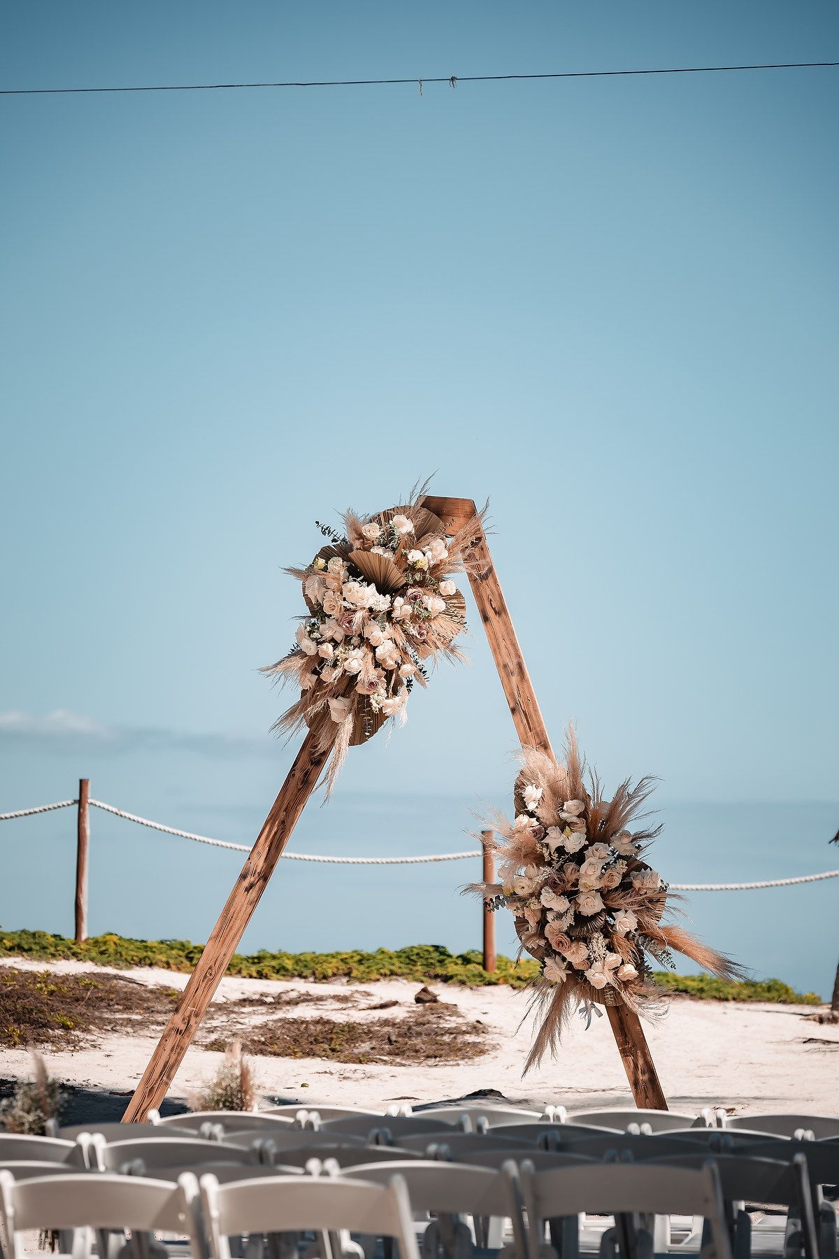 Wooden arch decorated with blush and white flowers stands on a beach with white chairs set up for a ceremony.