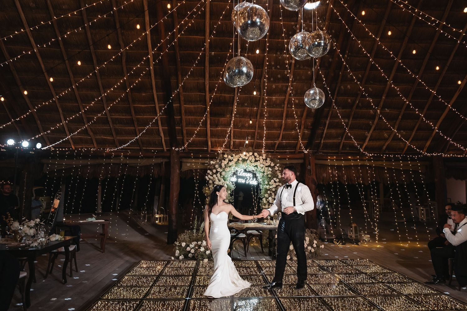 Bride and groom share first dance in a romantic barn venue adorned with fairy lights, disco balls, and a floral arch backdrop.