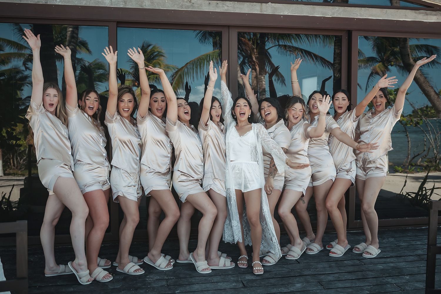 Bride and bridesmaids in matching white satin pajamas celebrate joyfully on a tropical resort deck with palm trees.