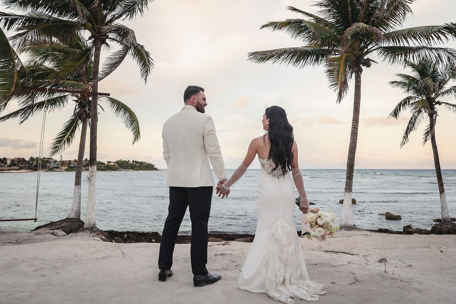 Bride and groom hold hands on a tropical beach at sunset with palm trees and ocean views.