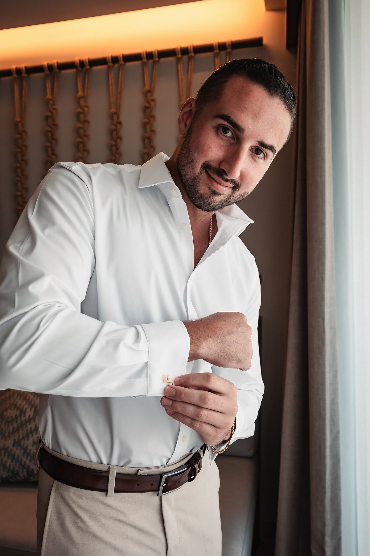 Smiling man in white dress shirt fastening cufflinks near a window in an elegant room.