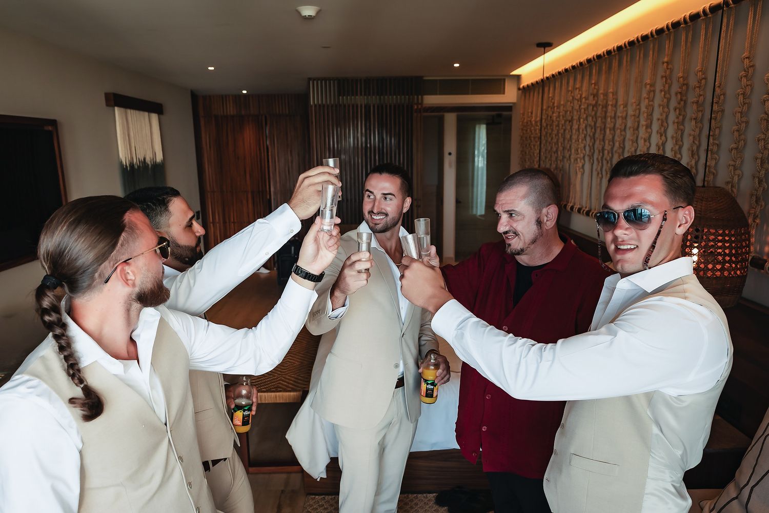 Five men in suits toasting with drinks at a wedding celebration in a hotel room.