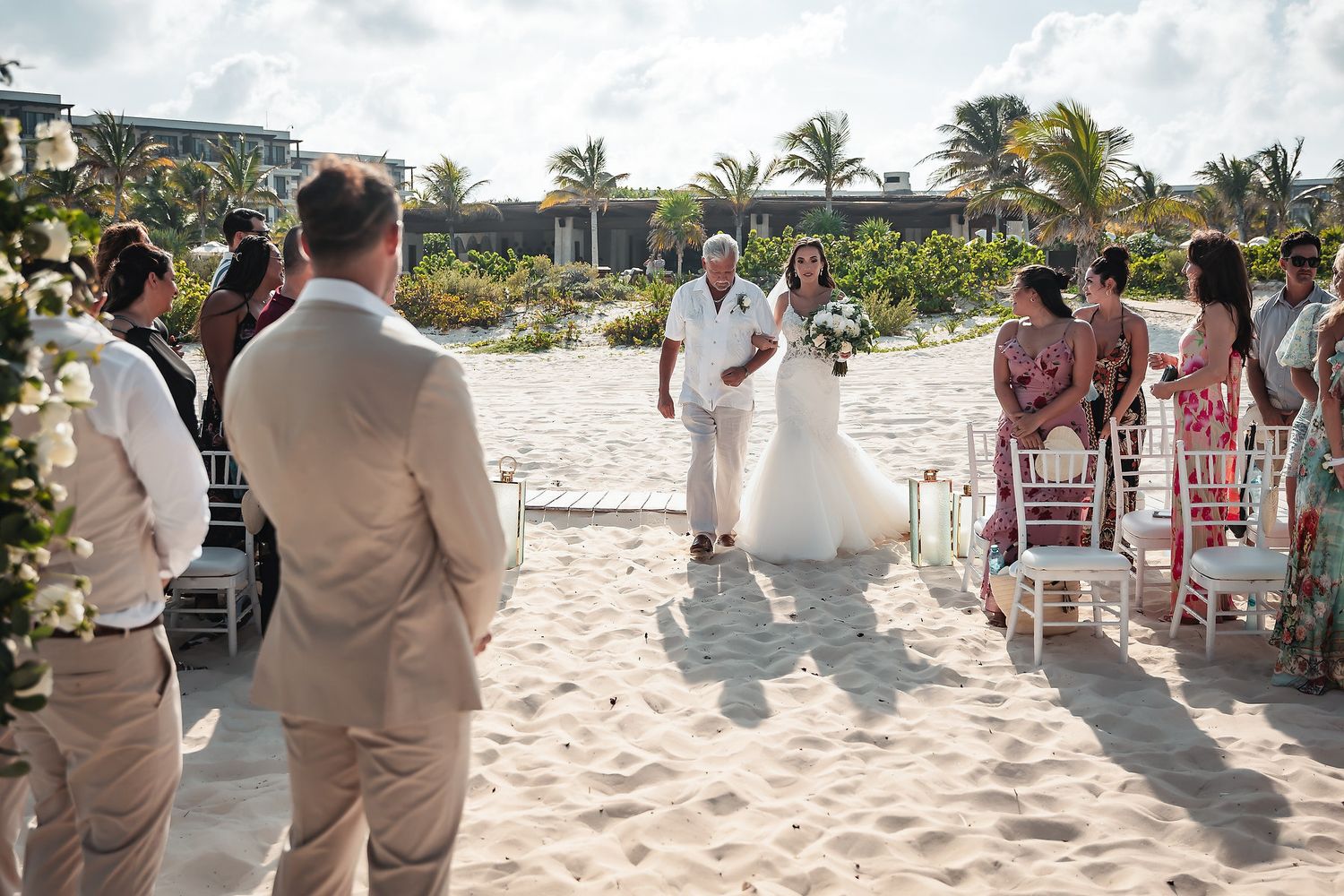 Bride walks down sandy beach aisle with officiant during an outdoor destination wedding ceremony.