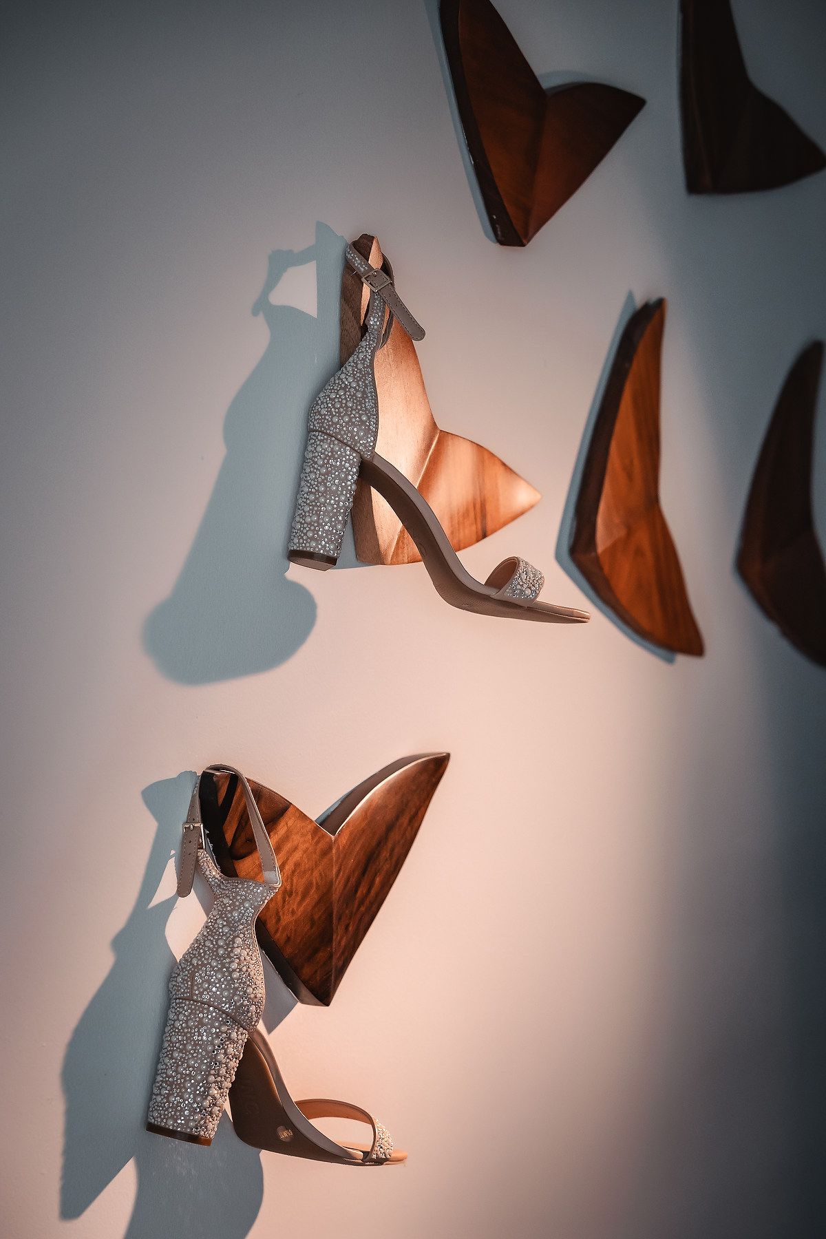 Glittery block heel sandals displayed on a white wall alongside abstract wooden sculptural shapes in warm lighting.