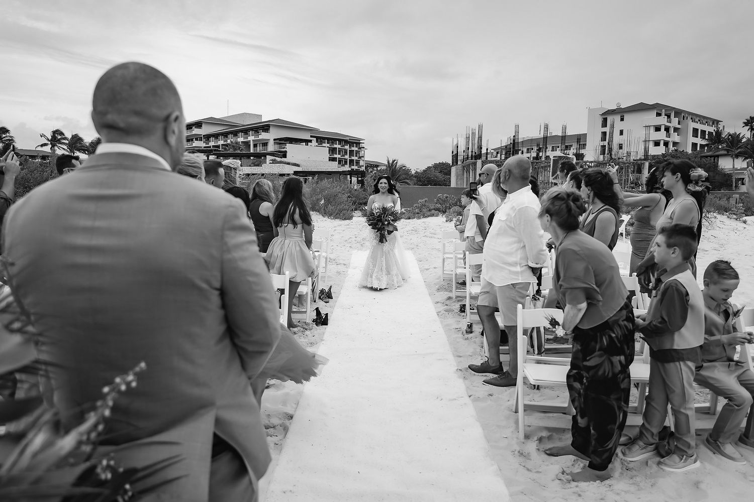 Black and white outdoor beach wedding ceremony with bride walking down aisle toward altar, guests seated on white chairs.