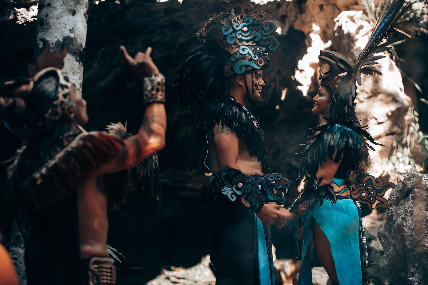 Performers in elaborate Mayan costumes with feathered headdresses and face paint gather in a cave setting.