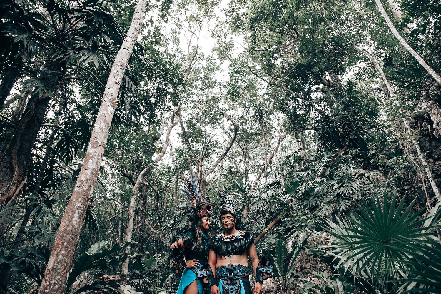Two performers in Mayan ceremonial costumes with feathered headdresses posing beneath tall jungle trees.