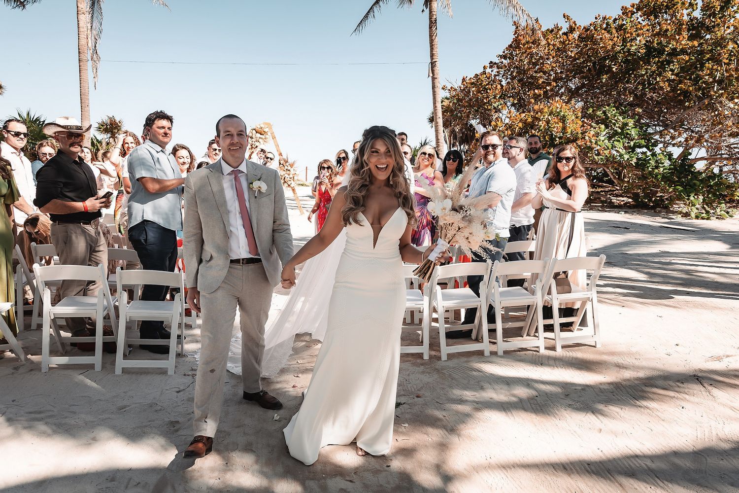 Bride and groom walk hand-in-hand down sandy beach wedding aisle, guests seated in white chairs under sunny sky.