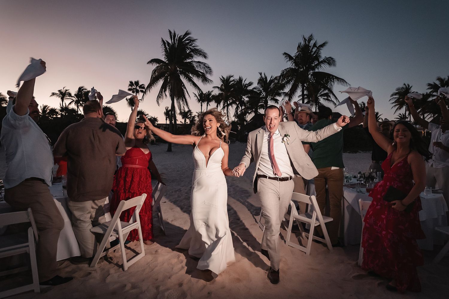 Happy couple exits beach wedding ceremony at dusk surrounded by cheering guests under palm trees.