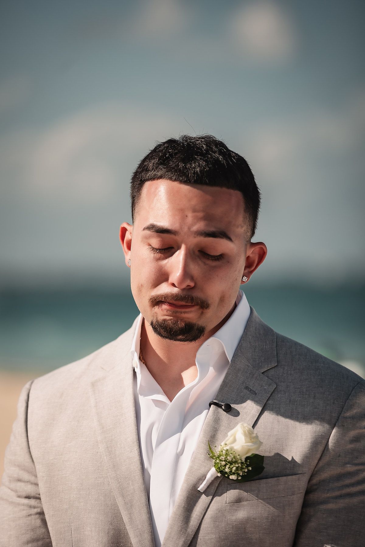 Emotional groom in gray suit with white boutonniere crying at beach wedding ceremony.