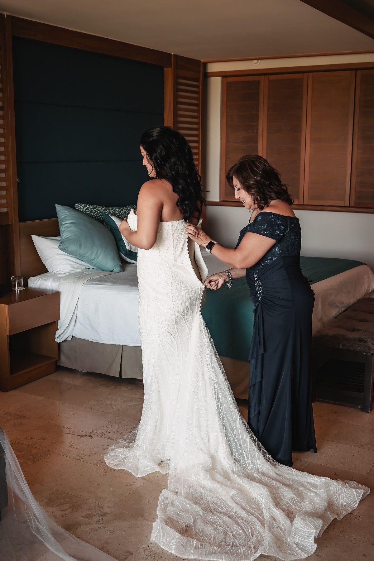 A woman in a navy gown buttons the back of a bride's white lace wedding dress in an elegant hotel room.