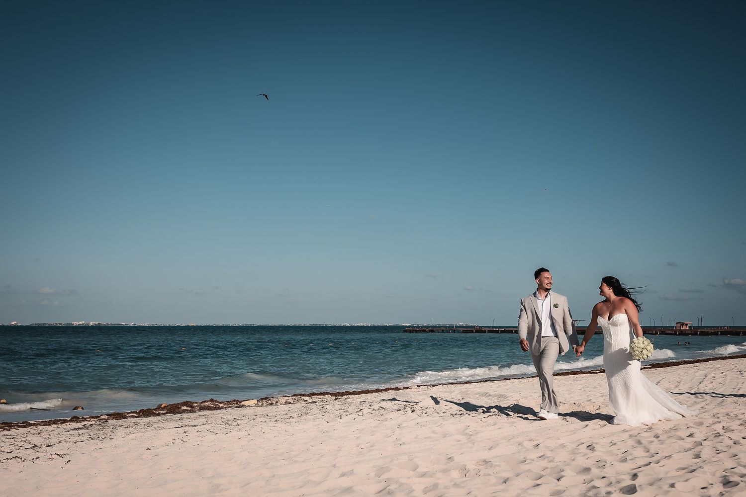 Bride and groom walking hand in hand on a white sandy beach with turquoise ocean waters on a sunny day.
