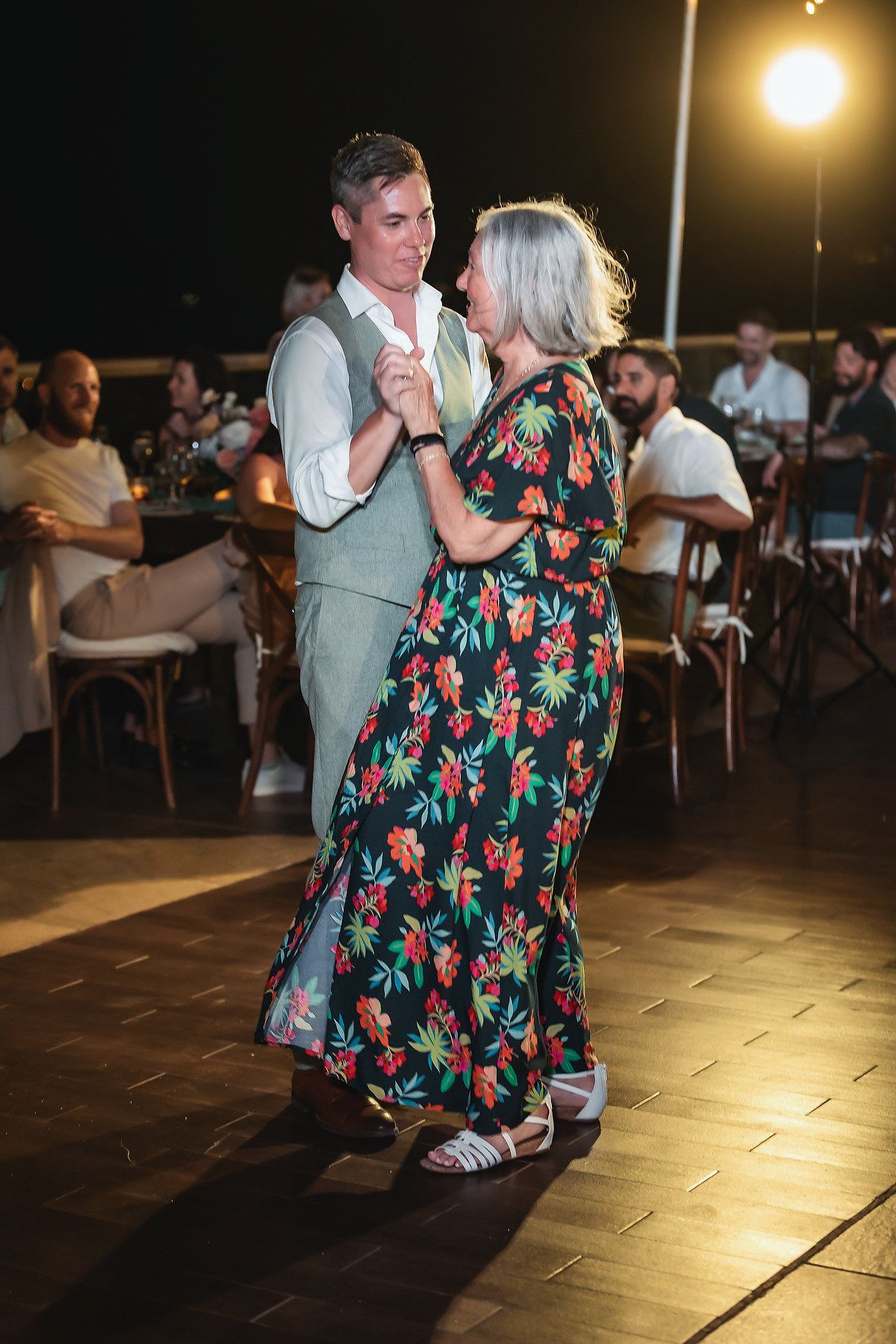 Couple dancing together at an outdoor evening event, woman in floral dress, man in vest, guests seated in background.