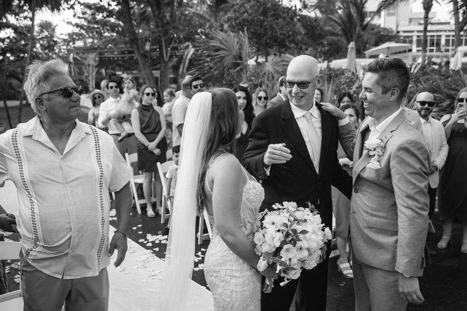Bride in white gown and veil holds bouquet as bald groom greets guests at outdoor wedding ceremony.